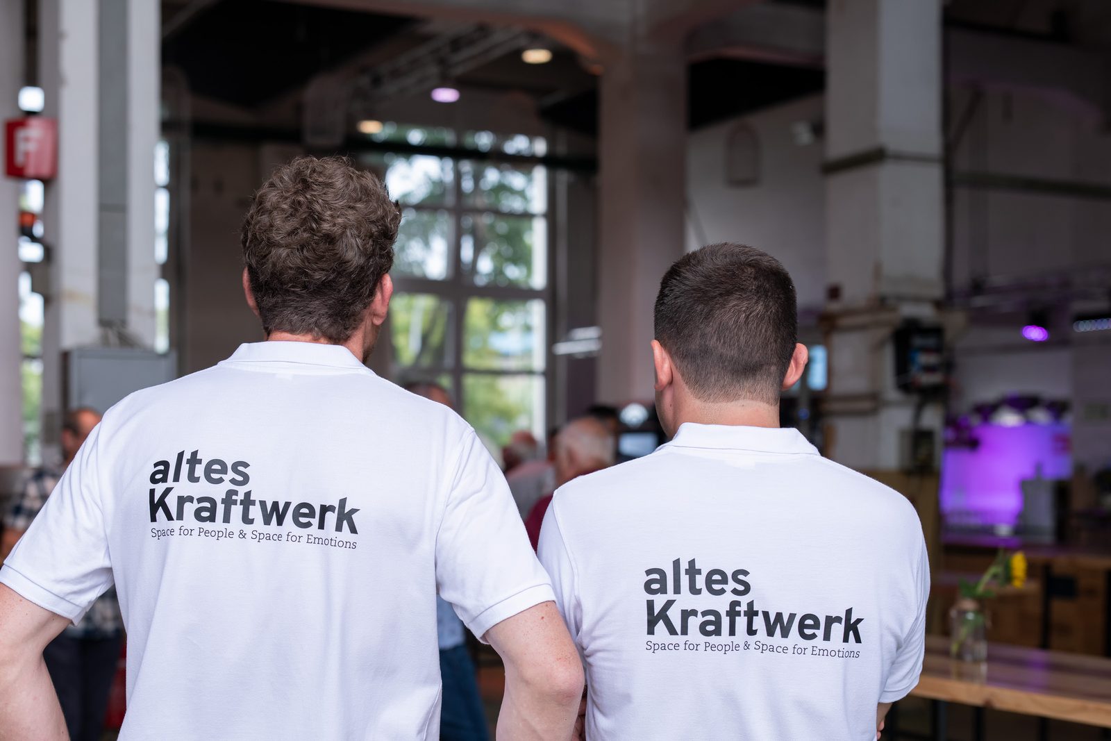 Zwei Mitarbeiter im Team-Poloshirt von hinten, Blick in die Eventhalle