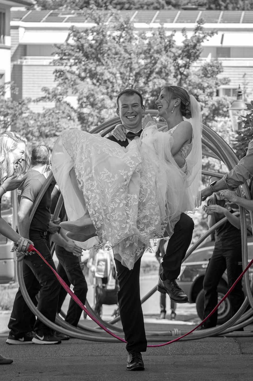 Bräutigam trägt die Braut lachend durch ein Spalier aus Feuerwehrschläuchen – Hochzeitsfotografie Region Basel