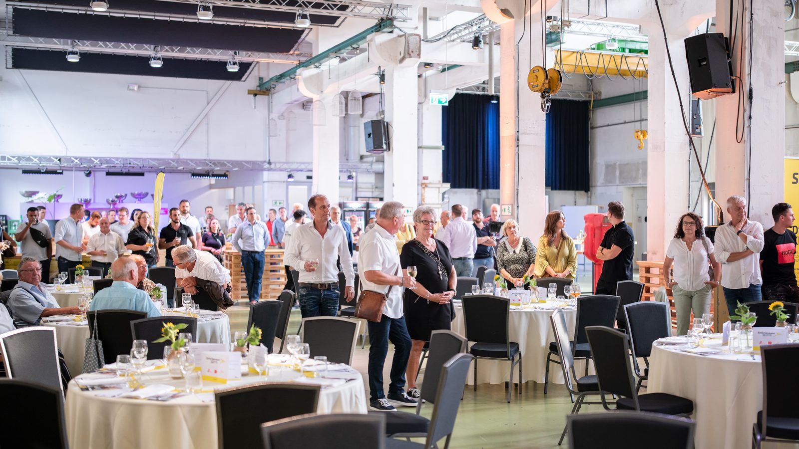 Gäste stehen zwischen festlich gedeckten Tischen in einer Industriehalle