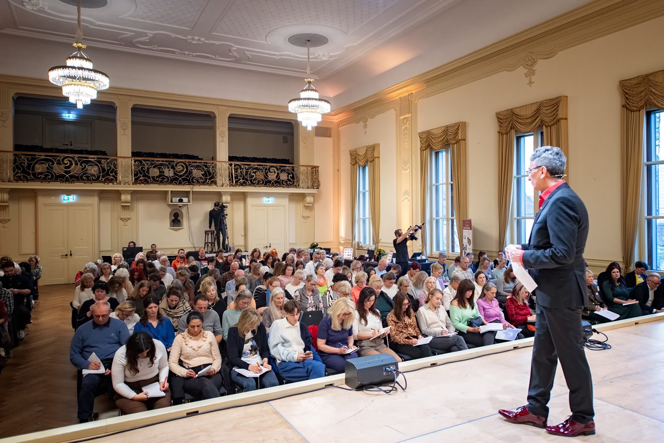 Referent auf der Bühne vor vollem Saal mit Kronleuchtern